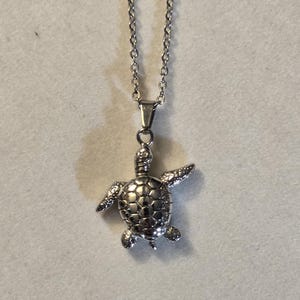 May include: A silver chain necklace with a silver turtle pendant. The turtle is detailed with a textured shell and has a realistic design.