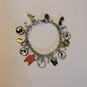Op de afbeelding: Een zilveren bedelarmband met diverse bedels, waaronder een rode jas, een muzieknoot, een wereldbol en een zwart-wit afbeelding van een persoon. De armband heeft een karabijnsluiting.