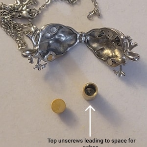 Op de afbeelding: Zilveren ketting met een scharnierende, opengewerkte hanger in de vorm van een vogel. De hanger heeft een klein, rond, goudkleurig bovenstuk dat losgeschroefd kan worden om een ​​ruimte voor as te onthullen.