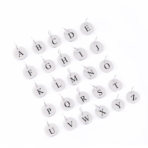 May include: A set of 26 silver-toned metal charms, each featuring a different letter of the alphabet. The charms are circular and have a small loop at the top for attaching to a necklace or bracelet.