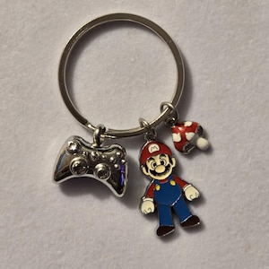May include: Silver keychain with three charms: a video game controller, a Mario figure, and a red and white mushroom. The Mario figure is wearing a red hat, blue overalls, and a red shirt.