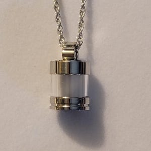 May include: A silver chain necklace with a cylindrical pendant. The pendant has a clear glass or plastic insert and a silver band around the top and bottom.