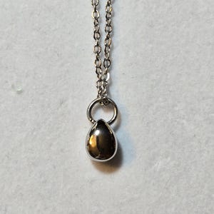 Könnte beinhalten: Eine silberfarbene Halskette mit einem tropfenförmigen Anhänger. Der Anhänger ist glatt und reflektierend und hängt an einem kleinen Ring, der mit einer feinen Kette verbunden ist. Die Kette besteht aus kleinen, verbundenen Gliedern.
