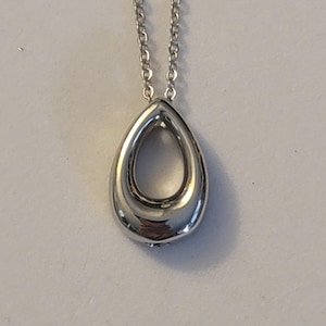 Puede incluir: Un collar de plata con un colgante en forma de lágrima con un centro hueco. La cadena es de plata y tiene un diseño de eslabones delicados.