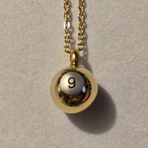 May include: A gold-colored necklace with a round pendant. The pendant is a gold-colored sphere with the number 9 in the center. The necklace chain is also gold-colored.