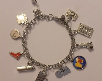 May include: A silver charm bracelet with various 80s themed charms, including a cassette tape, a roller skate, a can of hairspray, a boombox, a phone, a pair of Pac-Man ghosts, a red shoe, a camera, and a charm with the word "Duh" and the MTV logo.