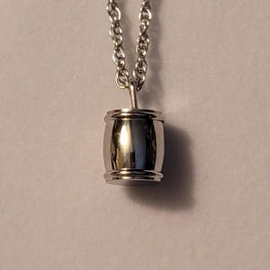 Può includere: Una collana con pendente color argento con un piccolo ciondolo a forma di botte. Il ciondolo ha una superficie lucida e riflettente ed è sospeso a una delicata catena. La collana è mostrata su uno sfondo neutro.
