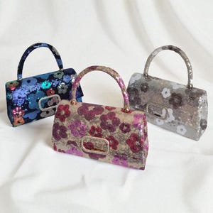 May include: Three sequined handbags with floral patterns. The bags have rectangular shapes, curved handles, and decorative clasps. The colours include blue, pink, and grey, with silver accents. The bags are on a white background.