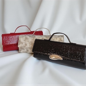 May include: Three patent leather clutch purses in red, silver, and black. Each has a matching handle and a flap closure. The black clutch features a gold-colored accent. The purses are displayed on a white, textured surface.