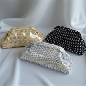 Peut inclure: Trois pochettes en maille de couleur or, argent et noir. Les sacs sont disposés sur un tissu blanc drapé. Le sac doré est à gauche, le sac argenté au centre et le sac noir à droite.