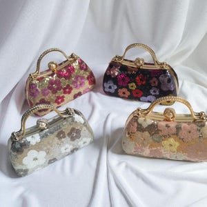 May include: Four sequined handbags with floral patterns and gold-tone handles. The bags feature various colors, including pink, gold, black, and silver, and are arranged on a white fabric background. Each bag has a clasp closure.