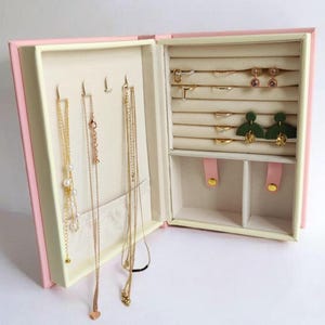 May include: A pink book-shaped jewellery box with a cream interior. The box has a section for necklaces, a section for rings, and a section for earrings. The box is open and the jewellery is visible.