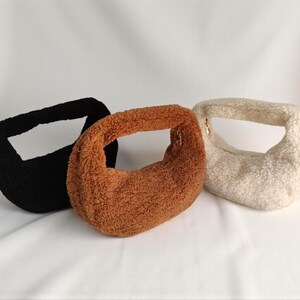 May include: Three fluffy, textured handbags in black, brown, and cream colors. Each bag has a rounded shape with a single handle and a gold-colored zipper. The bags are made of a soft, plush material.
