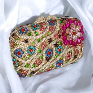 May include: A gold clutch purse with a floral design made of colorful rhinestones. The purse is decorated with pink, blue, green, and red rhinestones.