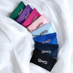 May include: A set of eight different colored satin clutches with a jeweled bow detail. The clutches are arranged in a row, with the darkest color at the bottom and the lightest color at the top.