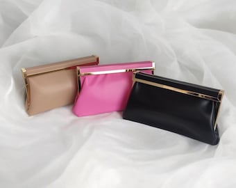 May include: Three clutch purses in a row, each with a rectangular shape and a gold-colored frame. The purses are in three colors: tan, hot pink, and black. The purses are resting on a white, draped fabric.