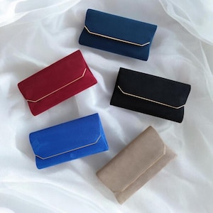 May include: Five different coloured clutch purses with gold trim. The purses are arranged on a white fabric background.