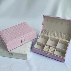 May include: Three jewellery boxes in shades of pink and white. The boxes are rectangular with a quilted pattern on the exterior. One box is open, revealing compartments and earring holders. The interior is lined with a light beige fabric.