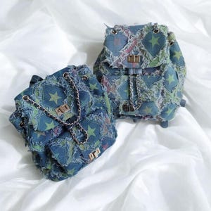 Op de afbeelding: Twee blauwe denim rugzakken met een sterrenpatroon en rafelige randen. De rugzakken hebben gouden kettingriemen en gouden hardware.
