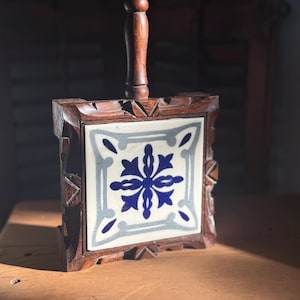 May include: A square trivet with a dark wood frame and handle. The center tile features a blue and white floral design. The trivet is designed to protect surfaces from hot cookware and adds a decorative touch to the kitchen.
