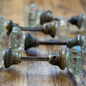 May include: A collection of vintage glass door knobs with a ribbed design. The knobs are clear with a slight green tint, and are attached to aged metal hardware. The knobs are arranged on a wooden surface.