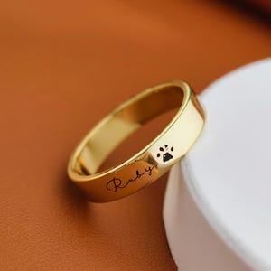 May include: A gold ring featuring a paw print and the name "Ruby" inscribed. The ring is displayed against a brown backdrop and a white surface. This is a piece of jewellery.