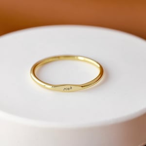May include: A gold ring with a rectangular nameplate. The ring is set on a white, circular surface against a brown background. The nameplate has an inscription in a foreign language.