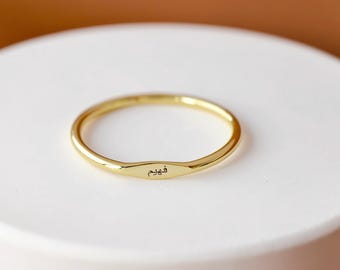 May include: A gold-colored ring with a rectangular nameplate. The ring is set on a white, circular surface against a brown background. The nameplate has an inscription in a foreign language.