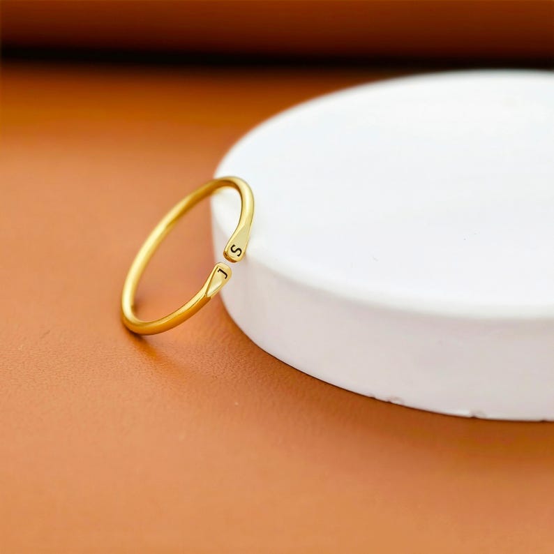 K&ouml;nnte beinhalten: Ein goldfarbener offener Ring mit einem d&uuml;nnen, glatten Band und polierter Oberfl&auml;che. Die beiden Enden des Rings sind leicht konisch zulaufend und jeweils mit einem einzelnen schwarzen Buchstaben, J und S, an der &Ouml;ffnung graviert.