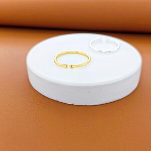 K&ouml;nnte beinhalten: Ein goldfarbener offener Ring mit den eingravierten Buchstaben J und S an den Enden, platziert neben einem schlichten silberfarbenen offenen Ring. Beide Ringe haben ein d&uuml;nnes, kreisf&ouml;rmiges Design und liegen auf einer glatten, runden wei&szlig;en Fl&auml;che.