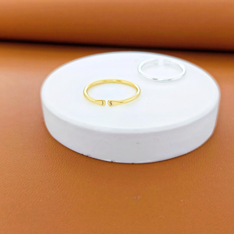 K&ouml;nnte beinhalten: Ein goldfarbener offener Ring mit den eingravierten Buchstaben J und S an den Enden, platziert neben einem schlichten silberfarbenen offenen Ring. Beide Ringe haben ein d&uuml;nnes, kreisf&ouml;rmiges Design und liegen auf einer glatten, runden wei&szlig;en Fl&auml;che.