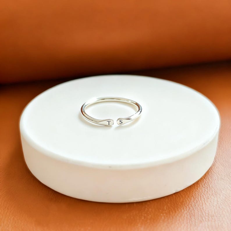 K&ouml;nnte beinhalten: Ein silberfarbener offener Ring mit einem d&uuml;nnen Band und zwei abgerundeten Enden. Jedes Ende ist mit einem einzelnen schwarzen Buchstaben versehen, wobei sich der Buchstabe T auf der linken und der Buchstabe S auf der rechten Seite befindet.