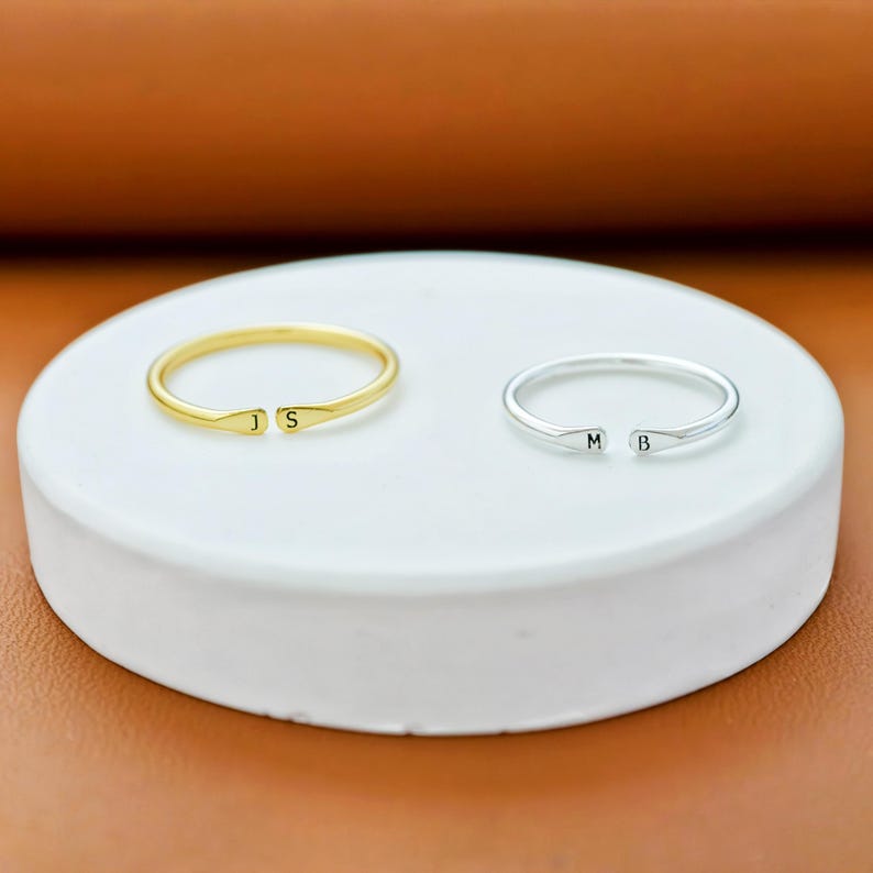 K&ouml;nnte beinhalten: Zwei offene Ringe, einer in Goldoptik und einer in Silberoptik. Der goldene Ring ist an den Enden mit den Buchstaben J und S graviert, w&auml;hrend der silberne Ring an den Enden mit den Buchstaben M und B graviert ist. Beide Ringe sind glatt poliert.