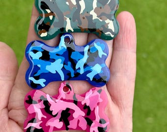May include: Three dog bone shaped tags with a camouflage pattern. The tags are green, blue, and pink with black accents.