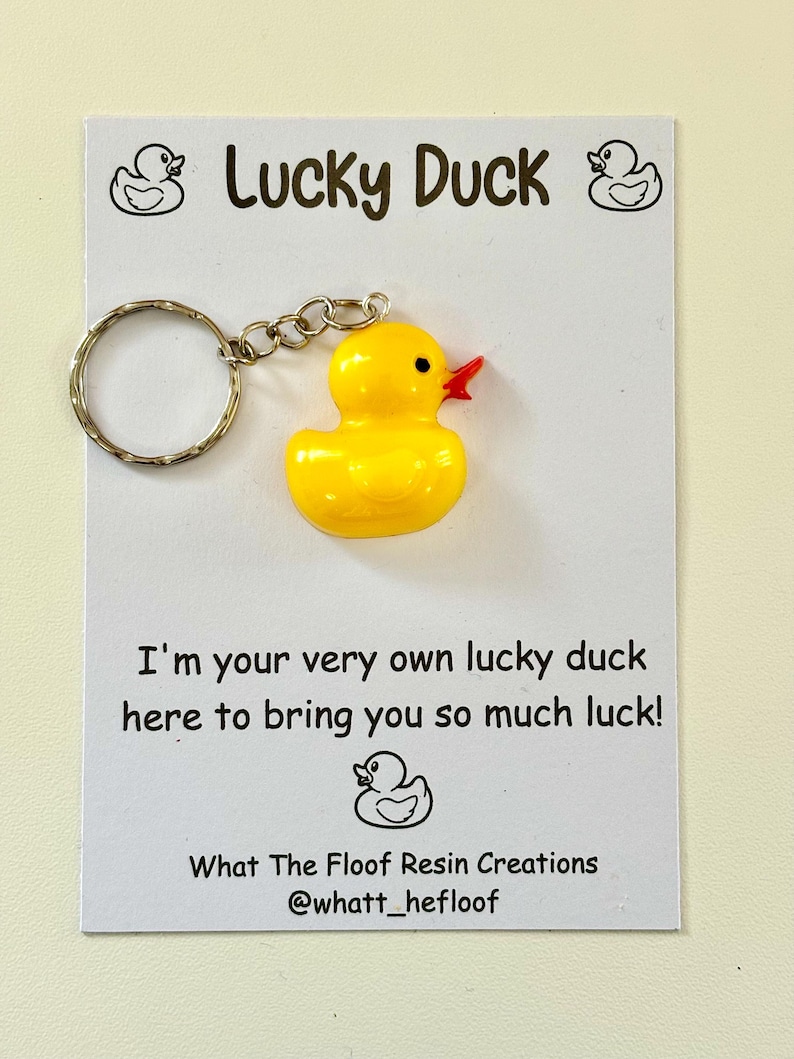 May include: A yellow rubber duck keyring with a red beak and a silver key ring. The keyring is attached to a white card with the text "Lucky Duck" and "I'm your very own lucky duck here to bring you so much luck!" and the text "What The Floof Resin Creations @whatt_hefloof".