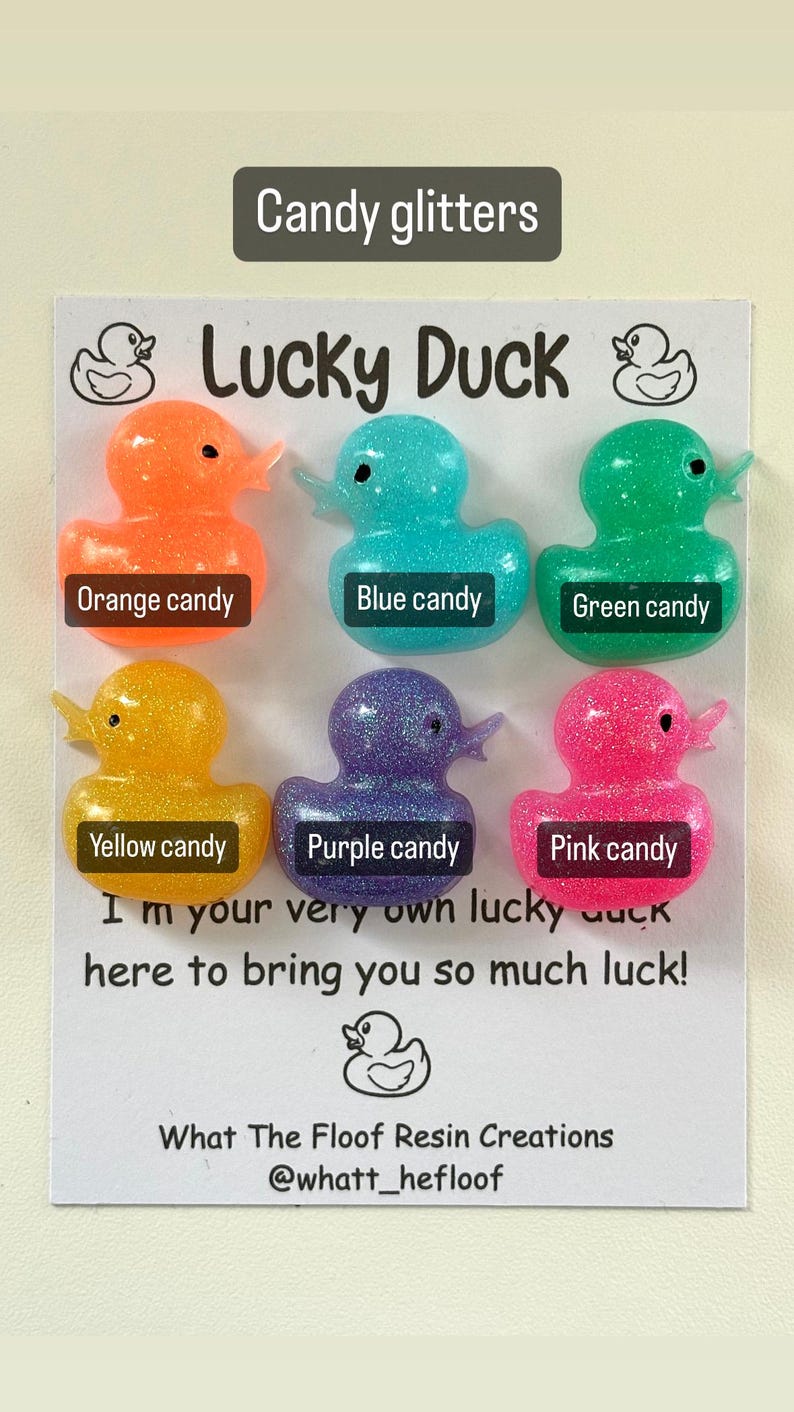 May include: Six small rubber duck figurines with glitter embedded in the plastic. The ducks are in the colours orange, blue, green, yellow, purple, and pink. The ducks are arranged in two rows of three. The text "Lucky Duck" is written in black letters on a white background. The text "I'm your very own lucky duck here to bring you so much luck!" is written in black letters on a white background. The text "What The Floof Resin Creations @whatt_hefloof" is written in black letters on a white background.