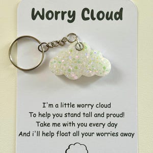 Pode incluir: Um chaveiro em forma de nuvem branca com glitter, com uma corrente e um anel prateados. O chaveiro tem um cartão anexado com o texto "Worry Cloud" e um poema: "Sou uma pequena nuvem de preocupação Para te ajudar a ficar alto e orgulhoso! Leve-me com você todos os dias E ajudarei a fazer todas as suas preocupações flutuarem."