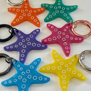 May include: A collection of colorful starfish keychains in orange, teal, purple, pink, blue, and yellow. Each starfish has a white dotted pattern and a metal keyring attachment. The keychains are a fun, beach-themed accessory.