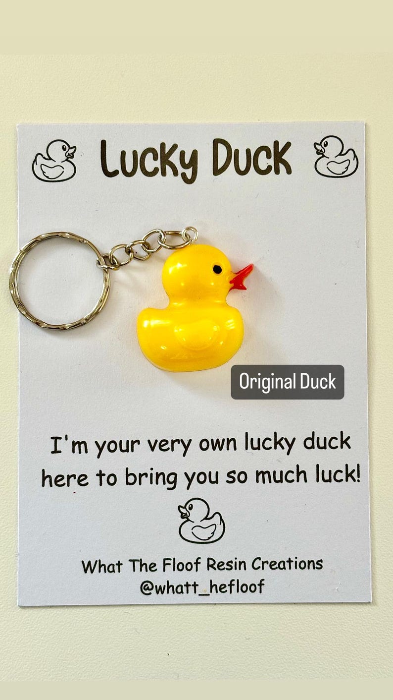 May include: A yellow rubber duck keyring with a red beak and black eyes. The keyring is attached to a silver key ring. The card reads "Lucky Duck" and "I'm your very own lucky duck here to bring you so much luck!" with a small rubber duck illustration at the bottom. The card also includes the text "What The Floof Resin Creations @whatt_hefloof".