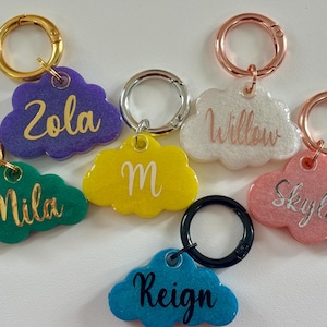 May include: A collection of cloud-shaped keyrings in shades of purple, green, yellow, white, pink, and blue. Each keyring features a name in cursive script. The clasps are gold, silver, rose gold, and black. Names include Zola, Mila, M, Willow, Skylar, and Reign.