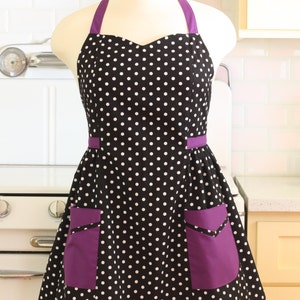 May include: A black and white polka dot apron with purple trim and two large purple pockets. The apron has a halter neck style.