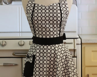 May include: A black and white polka dot apron with a black bow and a black pocket. The apron is on a mannequin in front of a white tiled wall and a vintage stove.