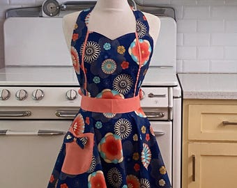 May include: A navy blue apron with a floral pattern featuring red, orange, and blue flowers. The apron has a peach-colored waistband, pocket, and trim. The apron is displayed on a mannequin in a kitchen setting.