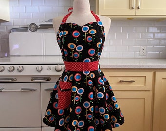 Puede incluir: Un delantal negro con una cintura de lunares rojos y un bolsillo rojo. El delantal tiene un estampado floral con flores azules, amarillas y rosas sobre un fondo negro. El delantal está en un maniquí blanco.