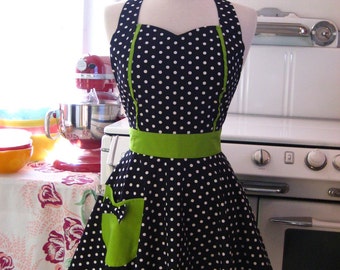 May include: A black and white polka dot apron with a green trim and a green pocket. The apron is on a mannequin in front of a white kitchen counter.