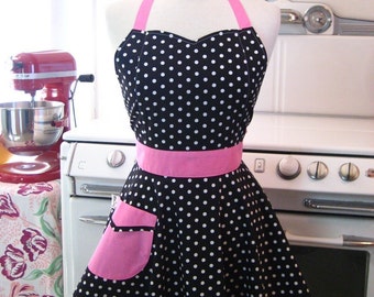 May include: A black and white polka dot apron with a pink waistband, pocket, and halter straps. The apron is displayed on a mannequin. A red stand mixer is visible in the background.