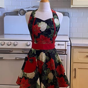May include: A festive apron with a black background, adorned with red poinsettias and white hydrangea blooms. The apron features a red waistband and pocket, and is displayed on a mannequin. The apron is designed for kitchen use.