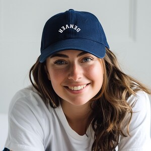 May include: A navy blue baseball cap with the word "DENVER" embroidered in white. The cap is worn by a person wearing a white t-shirt.