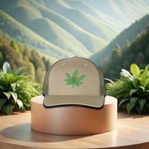 May include: A tan and green trucker hat with a smiling marijuana leaf design. The hat has a mesh back and a curved brim.