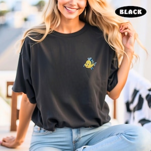 May include: Black t-shirt with a small embroidered yellow and blue cartoon fish. The word "BLACK" is in a black oval at the top right. The shirt is a crew neck and has short sleeves.